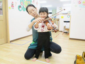 バイオリンを弾く保育園児と手伝う先生