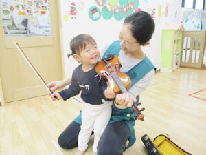 バイオリンを弾く保育園児と手伝う先生