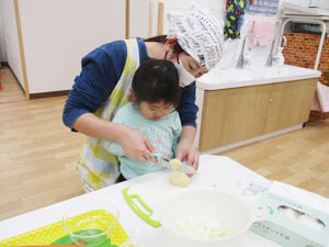 先生にお手伝いをしてもらいながら野菜を切る保育園児