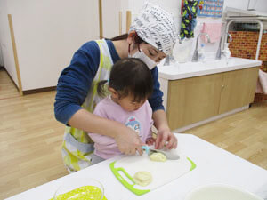 先生にお手伝いをしてもらいながら野菜を切る保育園児