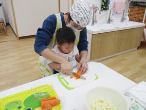 先生にお手伝いをしてもらいながら野菜を切る保育園児